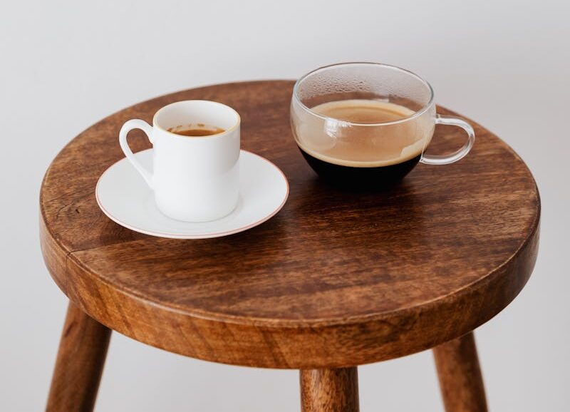 Fresh coffee in ceramic white cup and wide glass cup placed on round wooden table near white wall